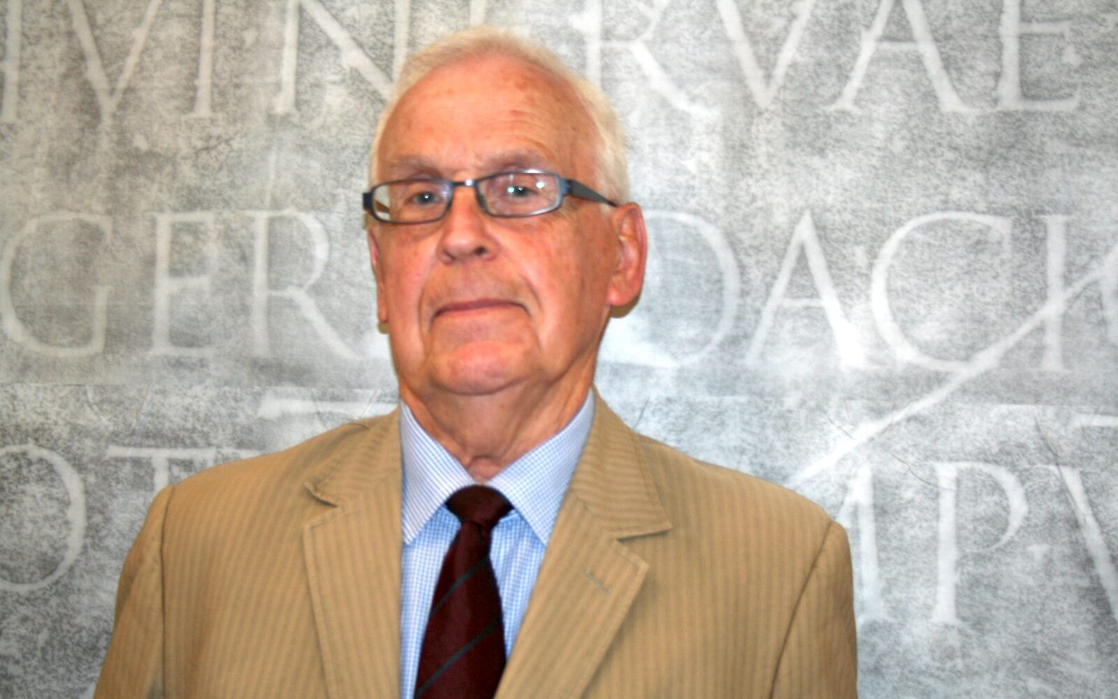 A headshot of a white-haired man in glasses wearing a dark tie, a blue-checked collared shirt, and a striped tan suit jacket. Behind him as a backdrop is a rubbing of a Latin inscription from Trajan's Column.