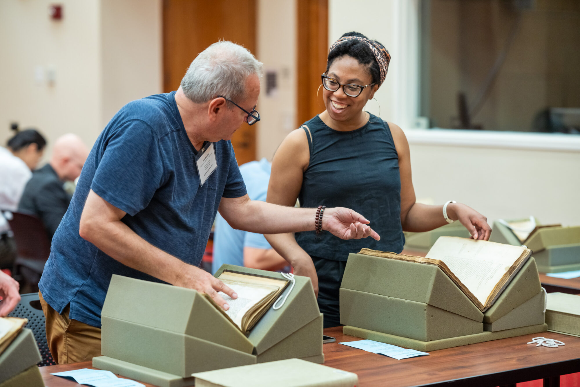 A man and a woman, both in glasses, stand side-by-side looking at books laid out on foam rests upon a long table. The man is pointing to something in the woman's book and she is smiling as he comments about it.