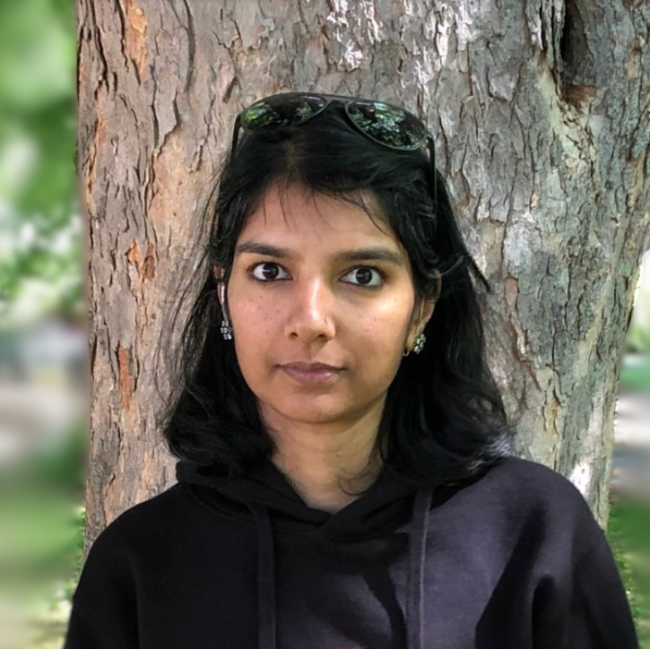 A dark-haired woman in a black hoodie stands in front of a tree trunk in tree-filled park.