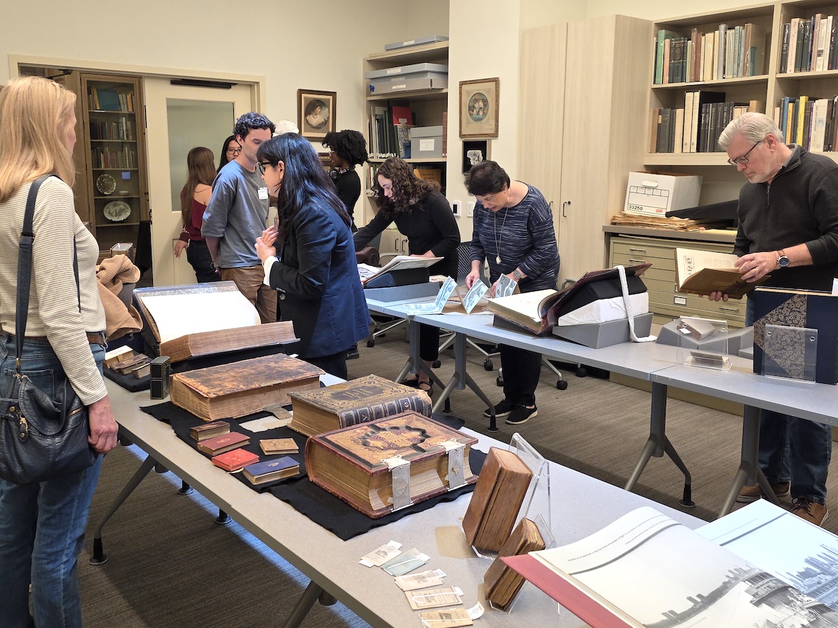 People spread out around two long tables placed parallel to one another, on which are laid out various rare books that the visitors are examining