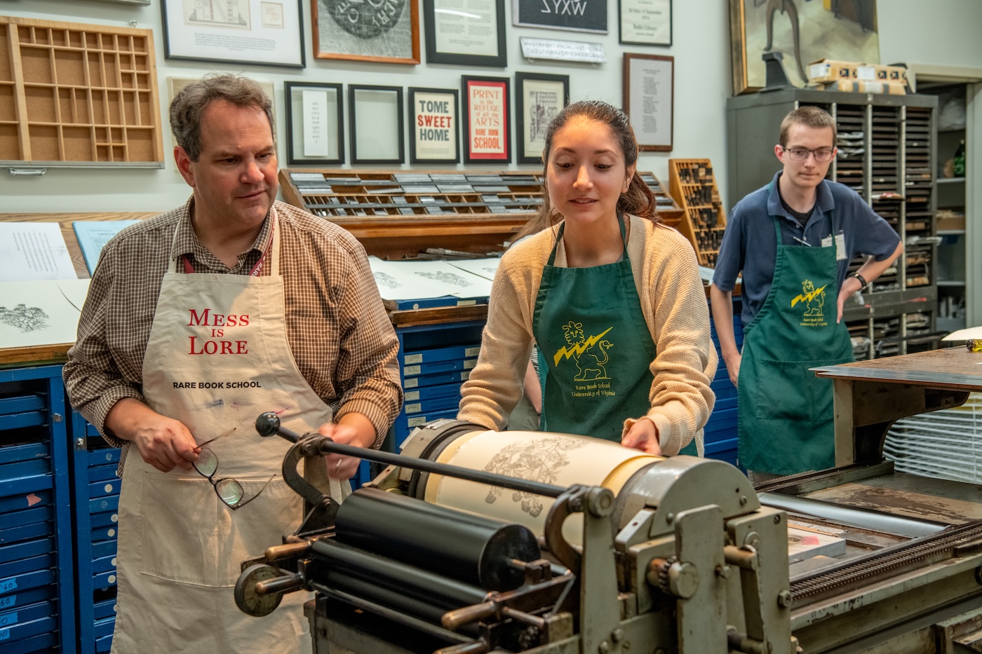 Three people in aprons stand in a printing office. The two men are looking on as the woman in the center turns the drum to print an image in black ink on a Vandercook press.