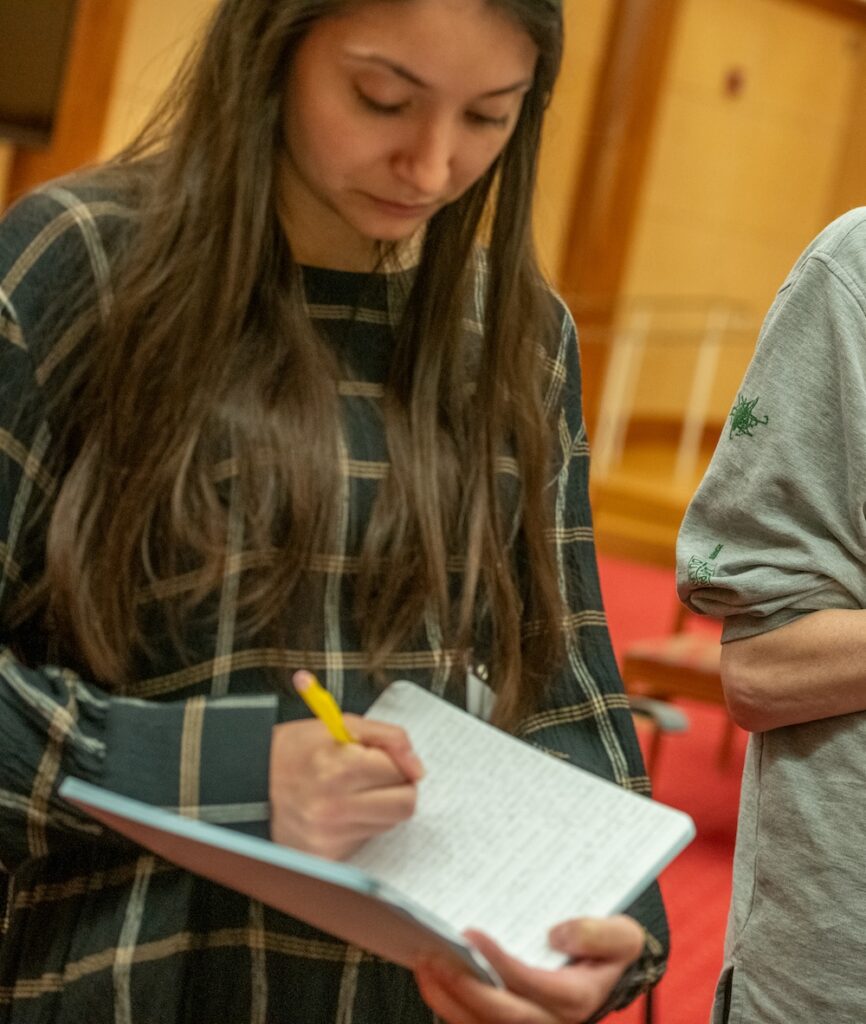 A woman in a green plaid dress stands writing in a notebook